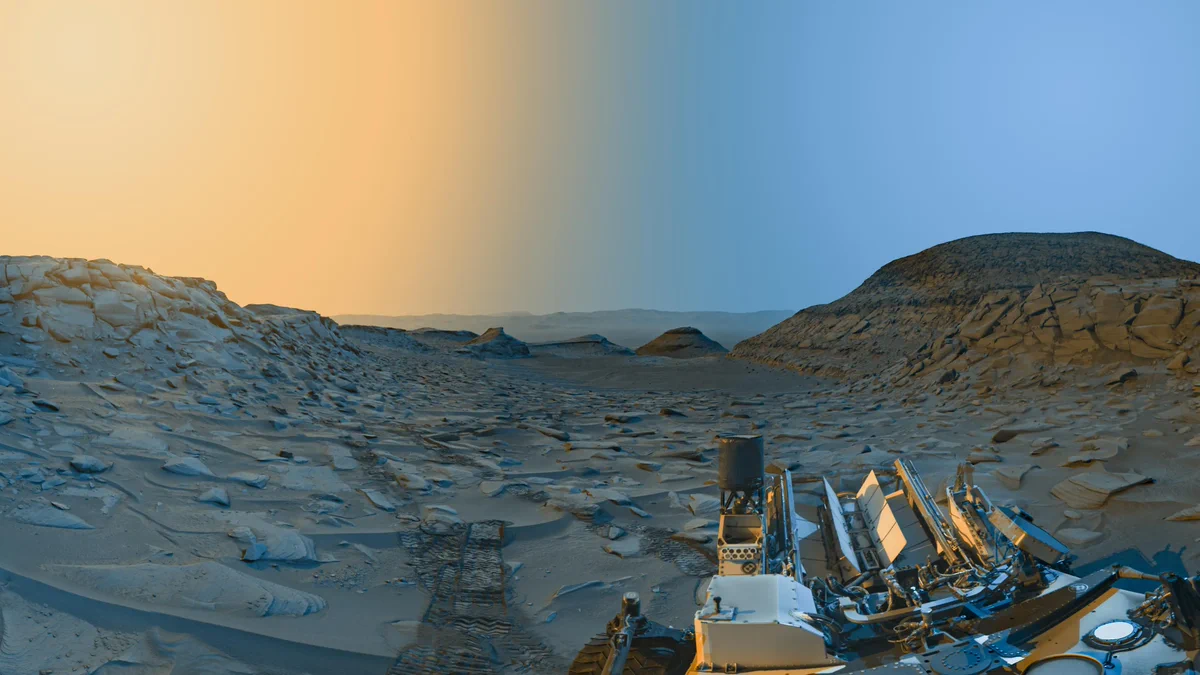 Two panoramas taken in the morning and afternoon on Mars are combined to create a color-enhanced landscape image. Both panoramas of the area, known as "Marker Band Valley," were taken by the Curiosity Mars rover's navigation cameras. The detailed photo shows yellow coloring in the sky to mark the afternoon hours and it transitions to a blue color to indicate the morning color. The taupe-colored landscape shows a large, rocky hill to the right and smaller rocky hills in the distance toward the center and out into the horizon. To the left is a rocky outcrop and as we get closer to the foreground the area shows rover tracks along the sandy floor. To the bottom-right corner, the backend of the off-white, dusty rover is shown. Part of the rover's wheel can be seen as well as three of its antennas. The Radiation Assessment Detector, or RAD instrument, appears as a white circle in the lower right portion on the back of the rover.