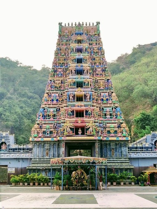 The image depicts the 8th Century Kanaka Durga/Sri Durga Malleswara Swamy Temple, located on the banks of the Krishna River in the Indrakeeladri Mountains of Vijayawada, Andhra Pradesh. The temple is a vibrant, multi-tiered structure with intricate carvings and colorful decorations. The architecture is traditional, with a towering gopuram (gateway tower) adorned with numerous statues of deities and mythological figures. The temple is set against a backdrop of lush green hills, enhancing its serene and sacred ambiance. The post text provides historical context, indicating the temple's long-standing presence and cultural significance.