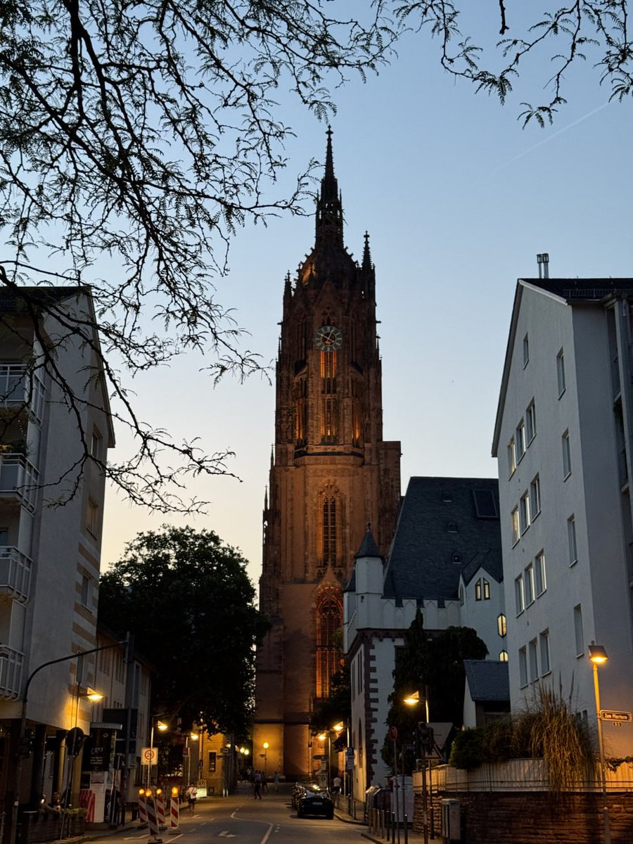 Evening view of a historic church tower in a quiet street, framed by trees and buildings.
