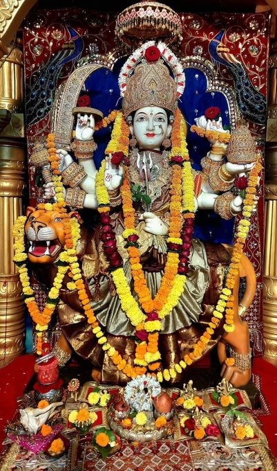 The image depicts a statue of the Hindu goddess Durga, known as Maa Durga or Maa Ambika, adorned with garlands of marigold flowers. The goddess is shown with eight arms, each holding different weapons and symbols, riding a lion, which aligns with the description in the post text as "अष्टभुजा धारी" (eight-armed). The setting is a richly decorated shrine or temple, with offerings placed at the base of the statue. The post text is a devotional hymn praising various aspects of the goddess, highlighting her power, compassion, and role in Hindu mythology. The image captures the essence of devotion and reverence towards the deity, reflecting the cultural and religious significance in Hinduism.