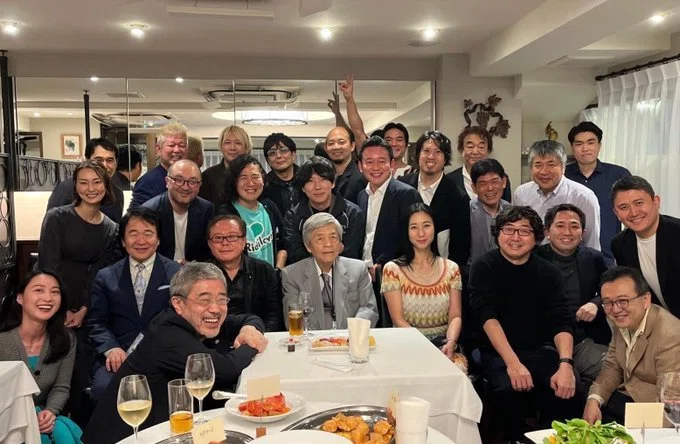 The image shows a large group of people gathered around tables in what appears to be a restaurant or dining setting. The group is dressed in formal and semi-formal attire, suggesting a professional or social gathering. The setting is well-lit with a warm ambiance, featuring framed pictures on the walls and a cozy atmosphere. The post text in Japanese translates to "Politics is all a farce," which implies a satirical or critical view of political gatherings, possibly suggesting that this group could be involved in political activities or discussions. There are no visible watermarks from social media platforms.