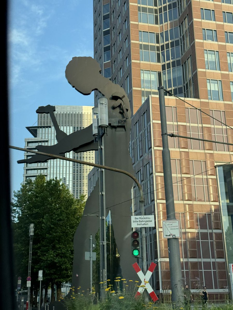 Large abstract sculpture in front of modern buildings, with a railway crossing sign and green traffic light, captured on a sunny day in an urban setting.