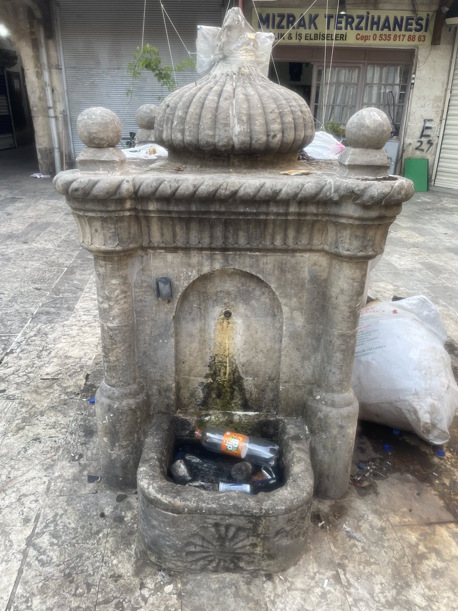 Eski Urfa’nın kaç yüzyıllık Mencek Hanı ve avlusu.Ortalık turist kaynıyor ve her yanı pislik götürüyor. Han’daki Çeşme’nin çöp kutusu olarak kullanılmasını önleyemeyen bir esnaf dayanamayıp kitabe yazıp asmış.