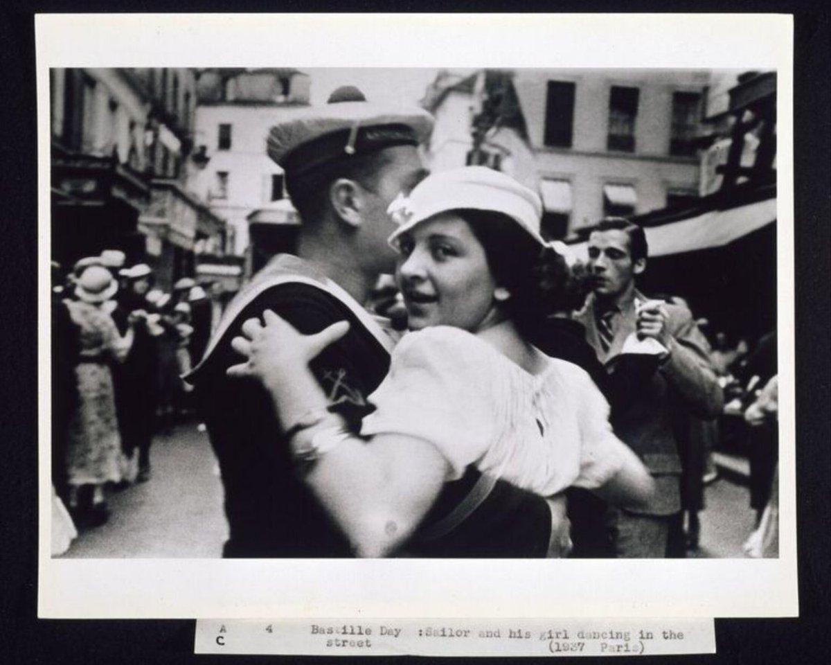 V_and_A's tweet image. A candid moment of joy, captured 88 years ago by Lucien Aigner. Titled Bastille Day, it shows a sailor and his companion dancing in the streets of Paris on July 14th, France’s national day, honouring the 1789 storming of the Bastille. What story do you see here?