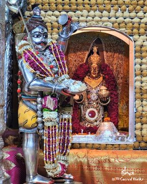 The first image depicts a golden statue of Goddess Annapurna, adorned with intricate jewelry and seated in a traditional pose, holding a bowl of food. The background is a rich red with ornate decorations, emphasizing her divine status. The text on the image reads "Goddess Annapurna of Kashi" and describes her role in making Lord Shiva realize the value of food. The second image shows a silver statue of Lord Shiva, adorned with garlands, receiving food from Goddess Annapurna, who is also depicted in this image. The setting is a temple, with a backdrop of red and gold, highlighting the sacredness of the scene. Both images are part of a post from Isha Sacred Walks, discussing the worship of Devi Annapurna in Kashi and the story where she made food vanish to teach Shiva a lesson about its importance.
