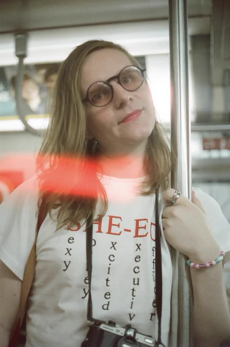 mb, a white trans woman, wearing a white t-shirt that reads "SHE-EO" across and "sexy, horney, excited, executive, officer" down under each letter. she’s wearing a film camera around her neck, circular blue glasses with rose tinted lenses, a red lip that’s faded a little, and a pink and blue bead bracelet as she grips the pole of an MTA train.