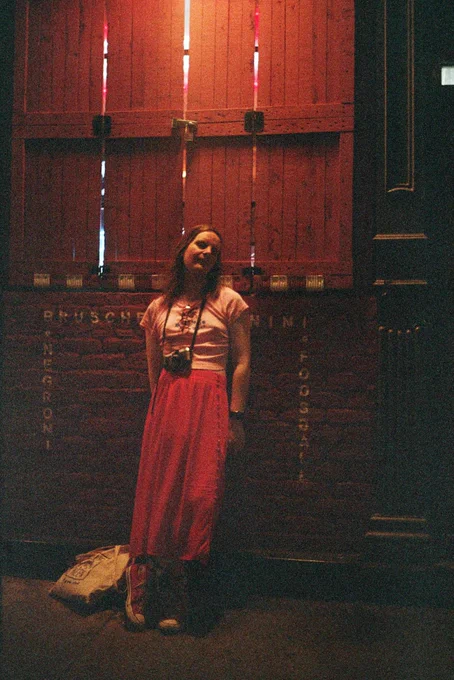 mb, a white trans woman, posing against a red wall late at night in new york city. shes’s wearing a pink crop top that says "tiny vibes", a long red skit, red Chuck Taylors, and a film camera. next to her sits a cream tote bag from Blue in Green.