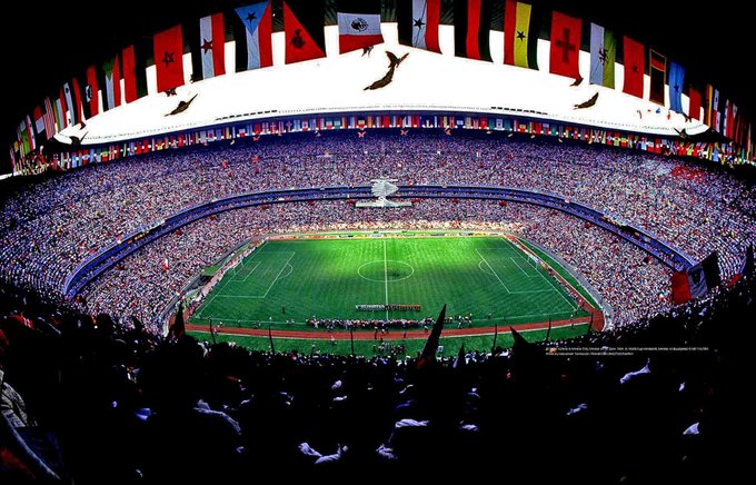 La imagen muestra un estadio de fútbol grande y abarrotado, probablemente el Estadio Azteca en la Ciudad de México, lleno de espectadores. El estadio está decorado con numerosas banderas nacionales de varios países, lo que indica un evento internacional. El contexto del texto de la publicación sugiere que esta imagen está relacionada con la inauguración planificada de la Copa Mundial de la FIFA 2026, que inicialmente se iba a celebrar en este lugar. Sin embargo, debido a los recientes disturbios en la Ciudad de México y las preocupaciones de seguridad planteadas por el gobierno canadiense, existe presión para trasladar el evento a Canadá. La imagen captura la grandeza y la anticipación de albergar un evento tan significativo, destacando la pérdida potencial para México si se reubica la inauguración.