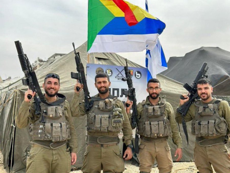 The image shows four men in military uniforms standing in front of a tent with mountains in the background. They are holding rifles and appear to be soldiers. Behind them, there are two flags: one is the Druze flag, characterized by its green, red, yellow, and white stripes, and the other is the flag of Israel. The context from the post text indicates that these men are Druze soldiers who are supportive of and protective towards Israel, highlighting a theme of unity and sacrifice.