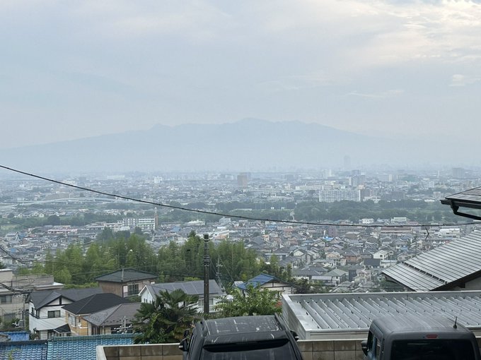 The image shows a distant view of Mount Akagi, which is faintly visible through the morning mist. In the foreground, there are rooftops of residential buildings and parked cars, indicating an urban setting. The post text mentions that there was a brief rain shower the previous evening, which refreshed the greenery around the house, and that Mount Akagi is visible, albeit faintly. The overall scene captures a serene morning with a blend of natural and urban elements.