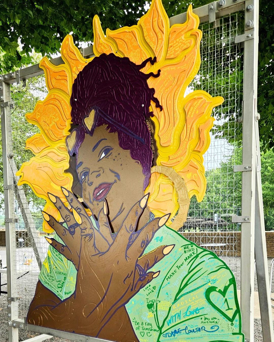 Colorful cut metal art by artist Dare Coulter, depicting a person with a radiant, sun-like sunflower halo behind their head, wearing a green shirt signed by Coulter and NCMA guests. The artwork is mounted on a metal fence outside East Building.
