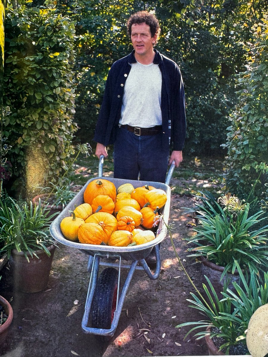 Monty Don wearing a white tshirt and jeans, pushing a wheelbarrow full of pumpkins in a garden.