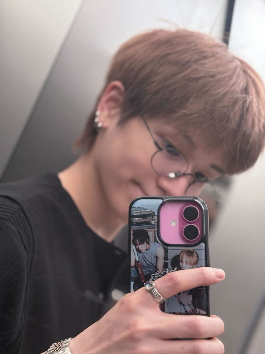 Round, wire-framed spectacles add an intellectual nuance, while multiple earrings and stacked silver rings introduce a whisper of rebellion. The phone case itself becomes a focal artifact, decorated with images that hint at artistic admiration.
