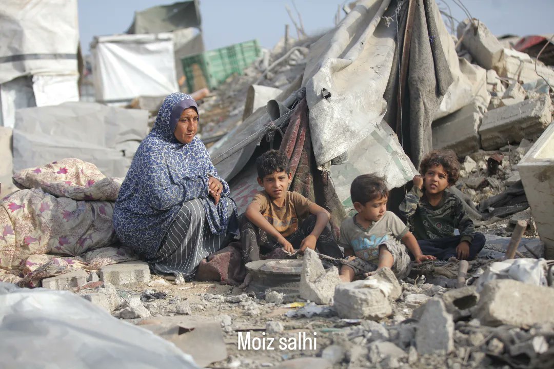 Timesofgaza's tweet image. A Palestinian family sits amid the rubble of a destroyed camp, as attacks continue in Gaza despite ongoing ceasefire negotiations.