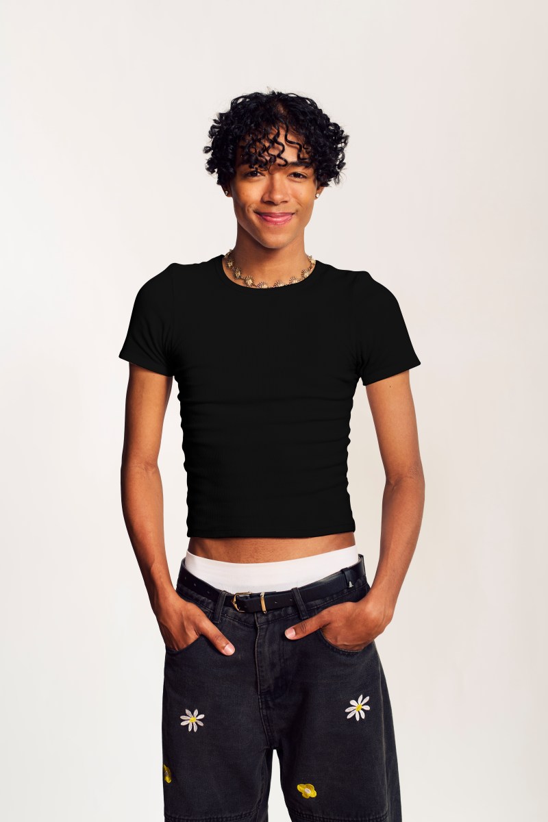 The image features Kauê Penna, an 18-year-old Brazilian member of the pop band Santos Bravos, as indicated by the post text. He is wearing a black cropped t-shirt and high-waisted jeans with embroidered daisies. The setting is a plain white background, which keeps the focus on his attire and expression. Kauê Penna is smiling, giving a friendly and approachable vibe. The post text provides his name, age, nationality, and Instagram handle, linking him to the band Santos Bravos, which is associated with HYBE, a major entertainment company.