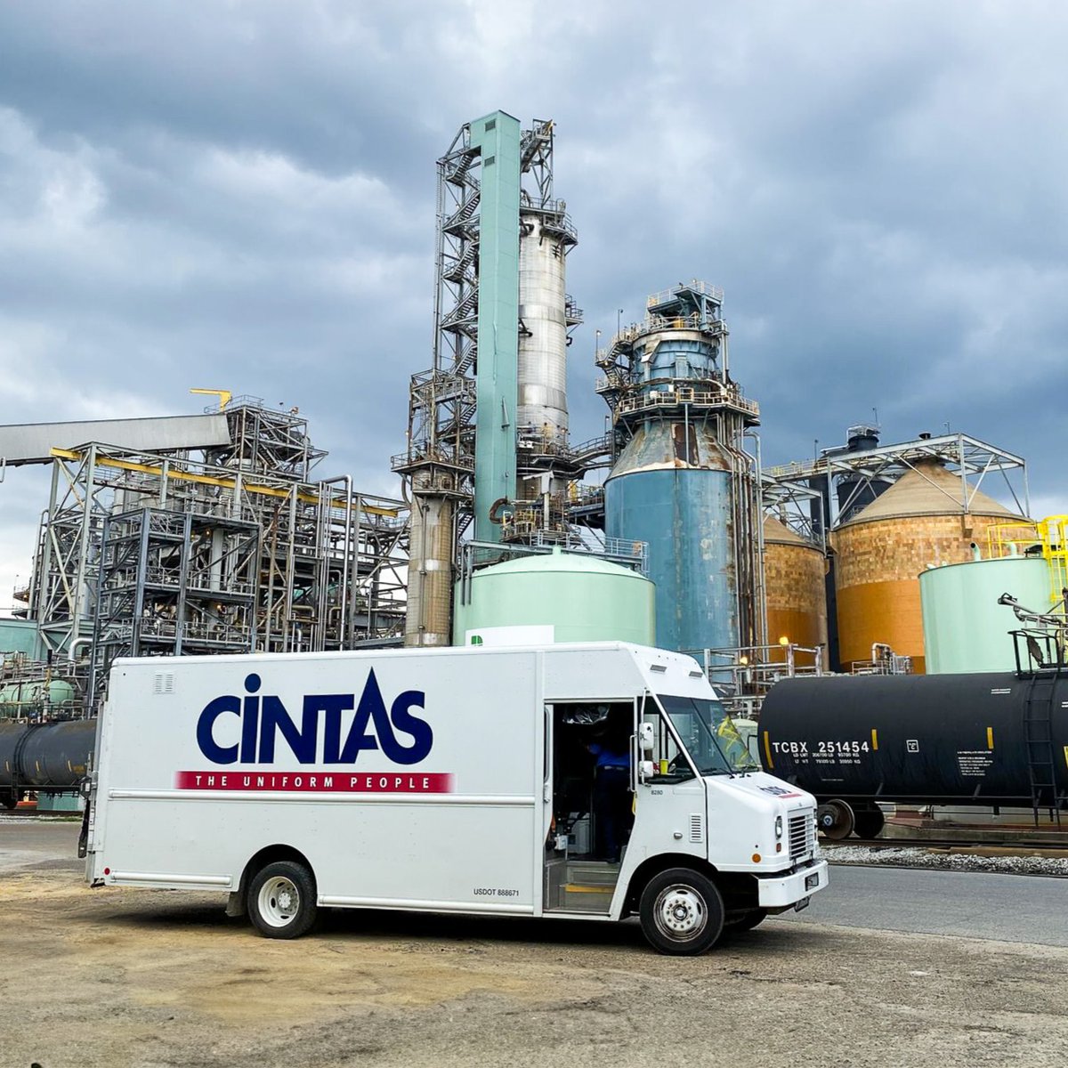 A Cintas delivery van parked in front of an industrial facility with towers, tanks, and pipelines under a cloudy sky.