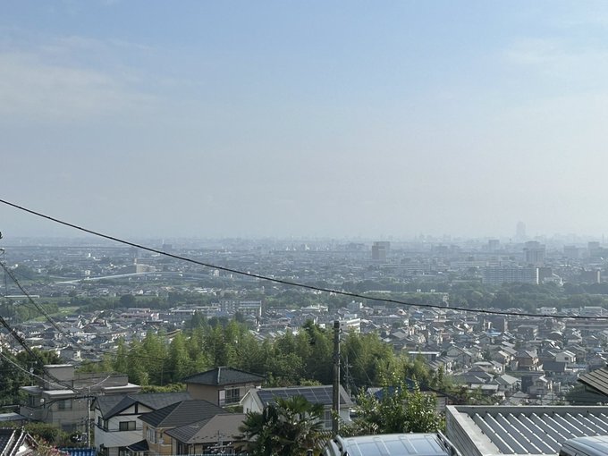The image shows a panoramic view of a cityscape with a clear blue sky above. In the distance, the silhouette of Mount Akagi is faintly visible, as mentioned in the post text. The foreground includes residential buildings and power lines, indicating an urban setting. The post text notes that the sky has cleared up, and while Mount Akagi is almost visible, it is not fully clear, adding context to the hazy appearance of the mountain in the background.