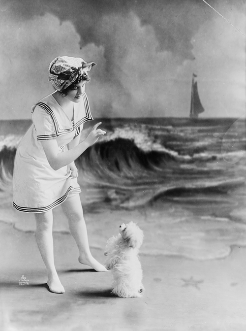 'Young woman in bathing suit with small dog in front of studio backdrop of beach'. Photographed in 1913.