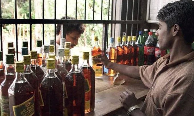 The image shows a man in a brown shirt handling bottles of alcohol, including whiskey and other spirits, on a wooden counter near a barred window, with another person visible in the background. Rows of liquor bottles with yellow caps are neatly arranged, suggesting a wine shop or liquor store setting. The post text from Telugu Scribe (@TeluguScribe) discusses the Telangana government under Revanth Reddy planning to expand wine shops to every village to boost alcohol sales and revenue, issuing preemptive notifications for upcoming elections, and adjusting liquor shop licenses and fees, indicating a policy-driven initiative.