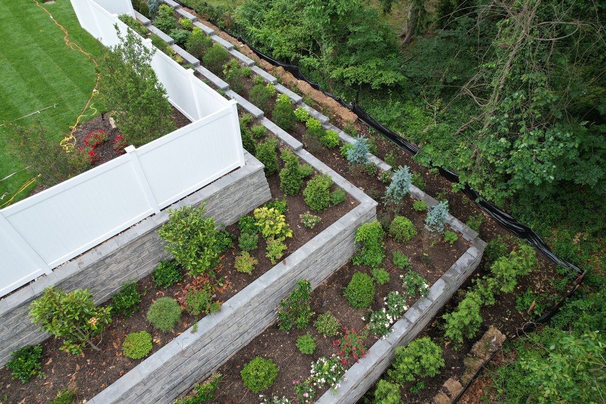 Tiered with purpose and designed for impact, this stunning retaining wall installation creates layers of beauty while maximizing space and structure. Each level is a canvas of greenery, bringing both function and curb appeal to life.Explore more ideas at CambridgePavers.com