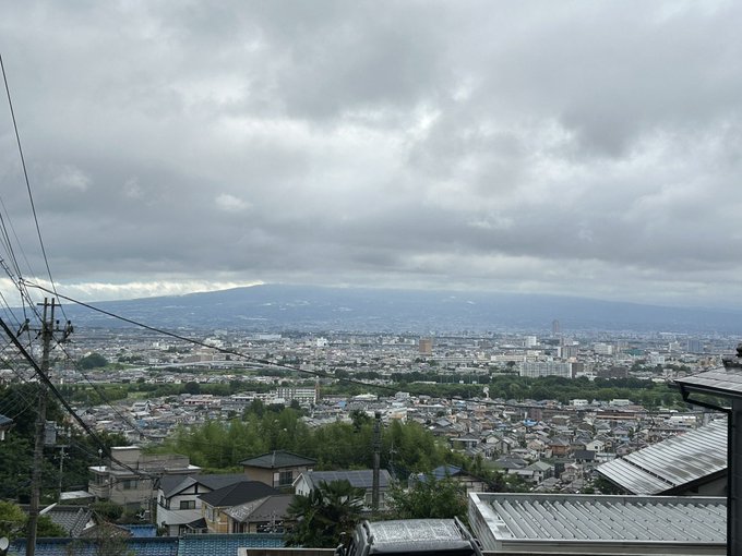 The image shows a panoramic view of a cityscape with a mountain, Akagi Mountain, partially visible in the distance under a cloudy sky. The foreground features residential rooftops, power lines, and greenery, suggesting a suburban area in Japan. The post text from きょうのあかぎ (@hs_akagi) indicates this is a rare sighting of the lower half of Akagi Mountain on July 17 at 7:10 AM, providing context for the cloudy weather and the mountain\'s visibility. No platform watermarks like Instagram, TikTok, or Xiaohongshu are present.