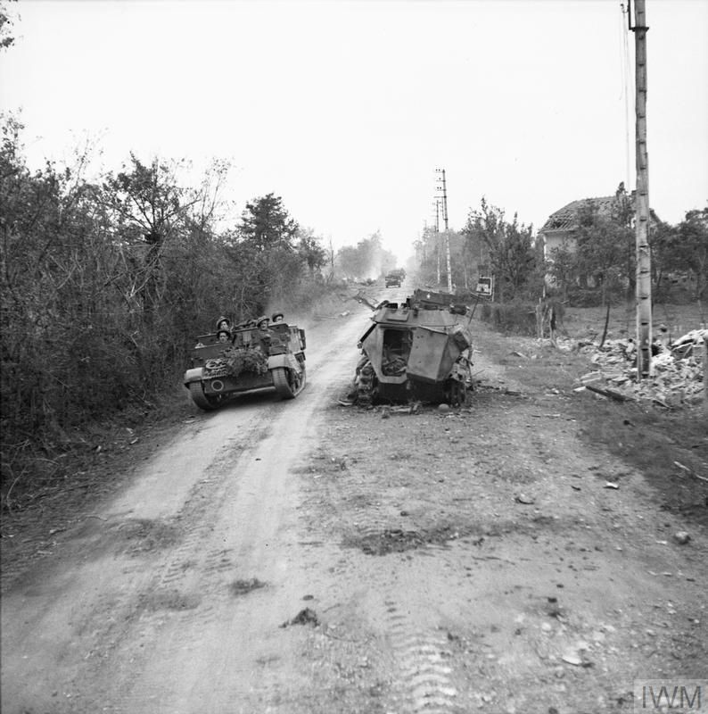 Normandy661944's tweet image. A Universal Carrier carrier passes a destroyed German half-track near Troarn during Op Goodwood, 19 July 1944. Ahead of the half-track, another carrier has turned off the road, perhaps abandoned. Further back, more carriers pass a smoking wreck.
IWM B 7686, Sgt Christie, AFPU
