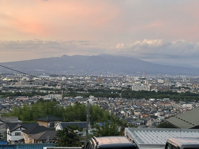 The image captures a serene evening view of a cityscape with Akagi Mountain visible in the background, as noted in the post text by きょうのあかぎ (@hs_akagi). The sky is painted with soft pink and orange hues of dusk, while scattered clouds add depth to the horizon. The foreground features residential rooftops, power lines, and parked vehicles, with the city lights beginning to twinkle as twilight sets in. The composition highlights the natural beauty of Akagi Mountain contrasting with the urban expanse below, creating a peaceful and picturesque scene. No platform watermarks like Instagram, TikTok, or Xiaohongshu are present.