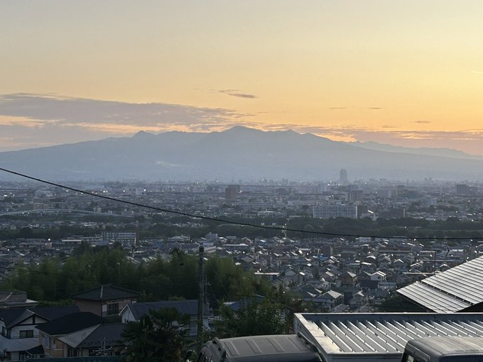 The image shows a serene sunrise view over a sprawling cityscape with the prominent Akagi Mountain range in the background, as mentioned in the post text by きょうのあかぎ (@hs_akagi). The scene features a mix of residential rooftops, trees, and power lines in the foreground, with the mountain\'s silhouette visible against a soft, golden sky. The post context reveals it was taken at 4:30 AM on July 20, with cicadas already chirping, and the author is at home drinking tea, unable to attend a planned soccer camp. No platform watermarks are present.