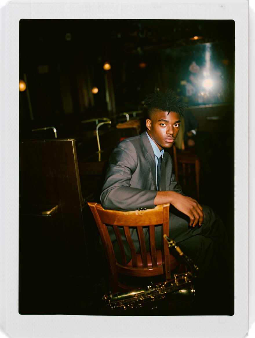Itsyopahadiboy's tweet image. Prompt: A vintage Polaroid snapshot of a young Black man in his early twenties, sporting a sharp, multi-pointed hairstyle. Dressed in a crisp grey suit jacket, light blue shirt, and dark tie, he sits at a dimly lit jazz club, a saxophone case resting beside him. The low-key…