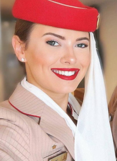 The image shows a smiling woman wearing an Emirates Airlines flight attendant uniform, featuring a beige jacket with red trim, a white scarf, and a red hat with a gold emblem. Her professional attire and cheerful expression suggest a positive representation of her role. The post text by @UltraDane indicates she is Danish and currently works for Emirates, aspiring to join Lufthansa, adding context to her career aspirations in aviation. No platform watermarks are visible.