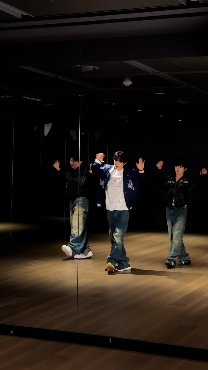 A person in a navy varsity jacket, white shirt, and baggy jeans in a dance studio, with their reflection seen in a wall mirror.