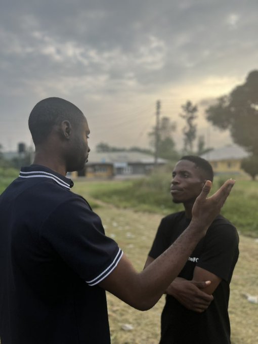 Two individuals discussing in an open field with a cloudy sky and buildings in the background