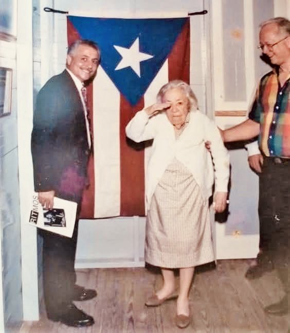 La Bandera de Puerto Rico desplegada, y que sirve de fondo para el saludo militar que la observamos aquí por Blanca Canales Torresola realizado solemnemente como Jefa de las Fuerzas Nacionalistas del 30 de octubre de 1950 en Jayuya, Puerto Rico. (1994)