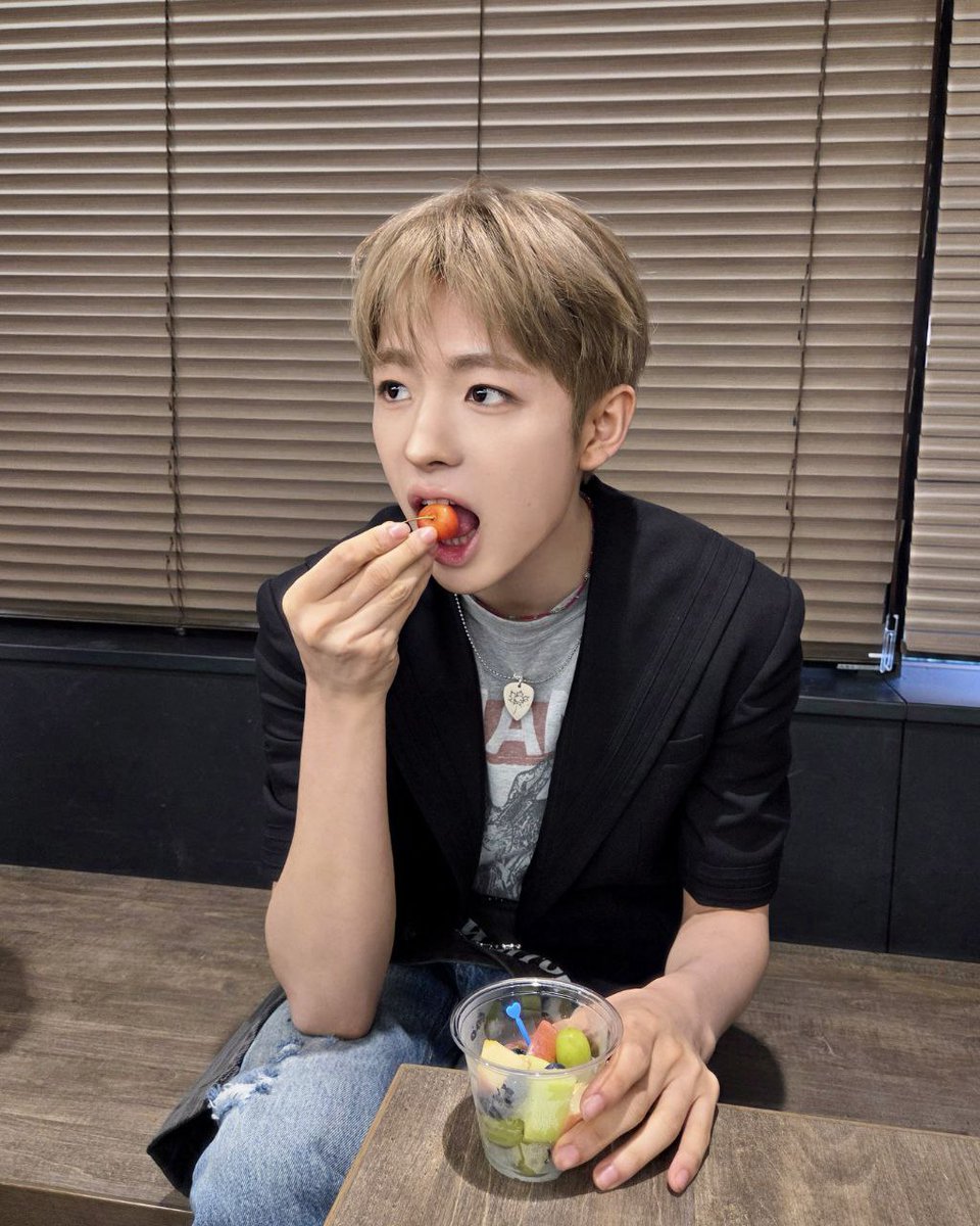 Black blazer over my graphic tee, ripped jeans on, holding a cup of fruit in one hand. I pick up a bright red cherry tomato and take a bite, the sweetness mixing with the freshness. The blinds behind me block out the world, making this feel like my own quiet break.