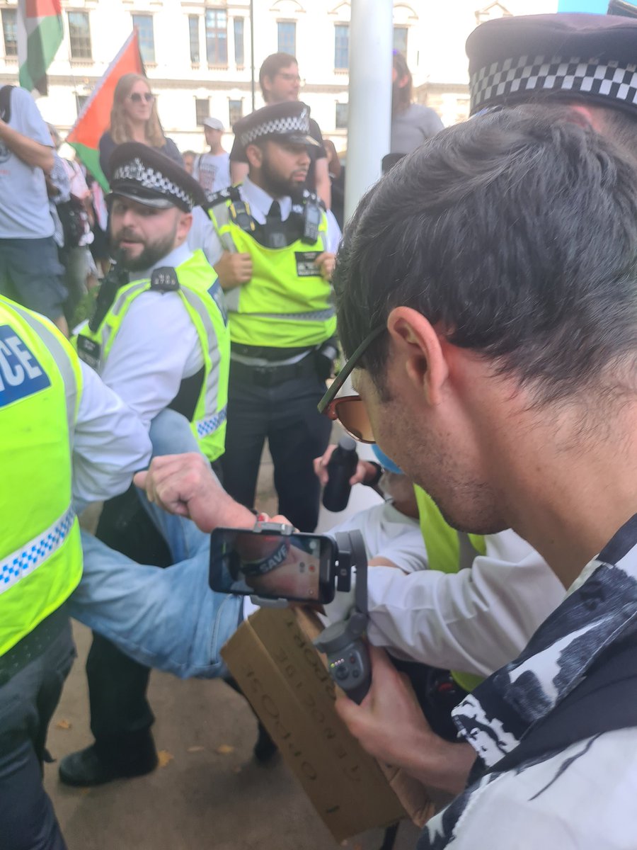 The protestor seen above, like many of those present, was admirably calm as she was surrounded by ten police officers, obscured from view, picked up, and carried away. Another man was carried away bodily.