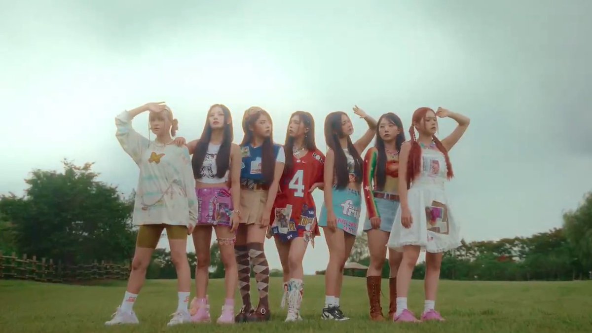 Seven young women in eclectic, colorful outfits stand in a line on a grassy field under a hazy sky, posing for a photo.