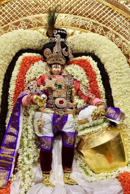 The image depicts Tirumala Sri Malayappan, a revered deity, adorned in elaborate traditional attire during the Chandra Prabhai Vahanam Brahmotsavam, as indicated by the post text from Tamil Brahmins. The deity is richly decorated with jewels, a peacock feather crown, and vibrant garments in red, white, and purple, holding a golden pot. Surrounded by garlands of white and orange flowers, the setting features an ornate golden backdrop, suggesting a temple festival. No platform watermarks are visible.