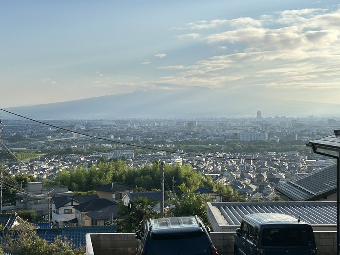 The image shows a panoramic view of a cityscape with a prominent mountain, Akagi Mountain, in the background, partially covered by clouds but clearly visible under a bright, clear sky. The foreground features residential rooftops, parked cars, and greenery, suggesting a suburban neighborhood. Power lines stretch across the scene, adding a typical urban touch. The post text from きょうのあかぎ (@hs_akagi) on August 1 at 5:58 AM indicates this is a morning view of Akagi Mountain and the county office, marking the start of August with a sense of renewal. No platform watermarks are present.