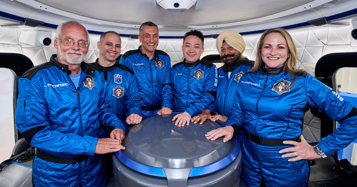 NS-34's six-person crew in Blue Origin flight suits smiling and standing inside a New Shepard training crew capsule. 