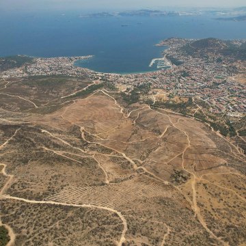 The images show an aerial view of a hilly, arid landscape in İzmir, Foça, with terraced, barren land and dirt paths, indicating areas cleared after a wildfire. Construction equipment like an excavator is visible, preparing the terrain for reforestation. The post text from Orman Genel Müdürlüğü (@OGMgovtr) explains that 234 hectares damaged by last year’s fire are being restored, with burned trees removed, studies completed, and land prepared for fall planting. The sea and a coastal town are visible in the background, adding context to the location. No platform watermarks are present.