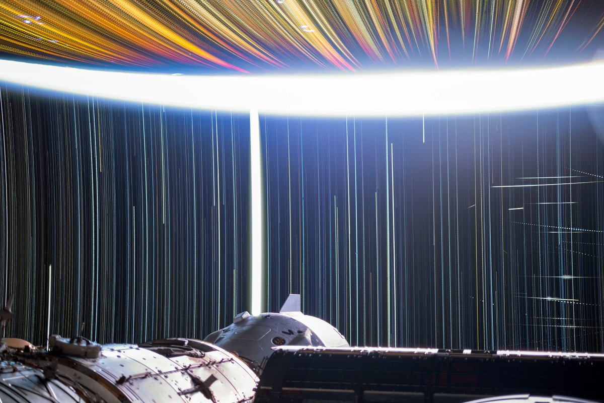 astro_Pettit's tweet image. Star trail exposure from the ISS, marked by the thick white path of the Moon, Starlink satellites flashing far right, golden city lights streaking across Earth, and arcing stars.