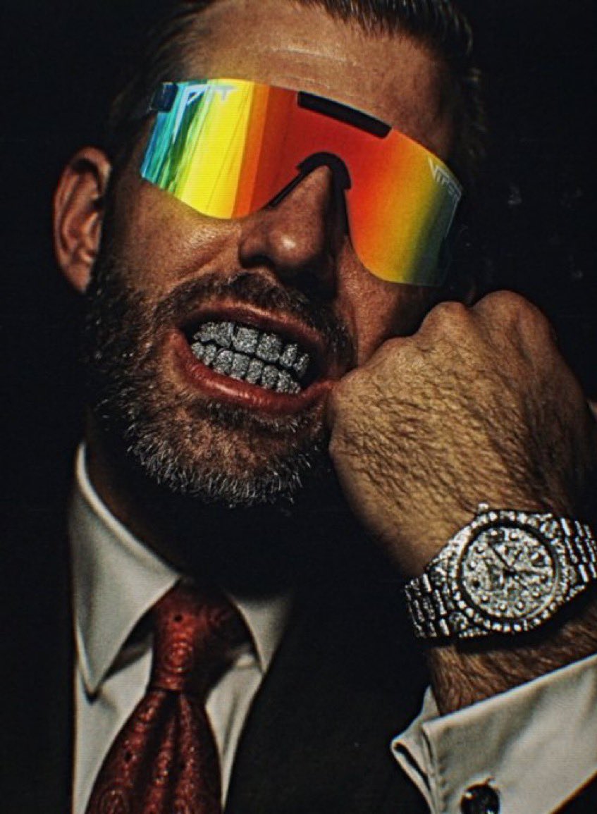 Man in suit with diamond grills and rainbow pit viper sunglasses posing confidently
