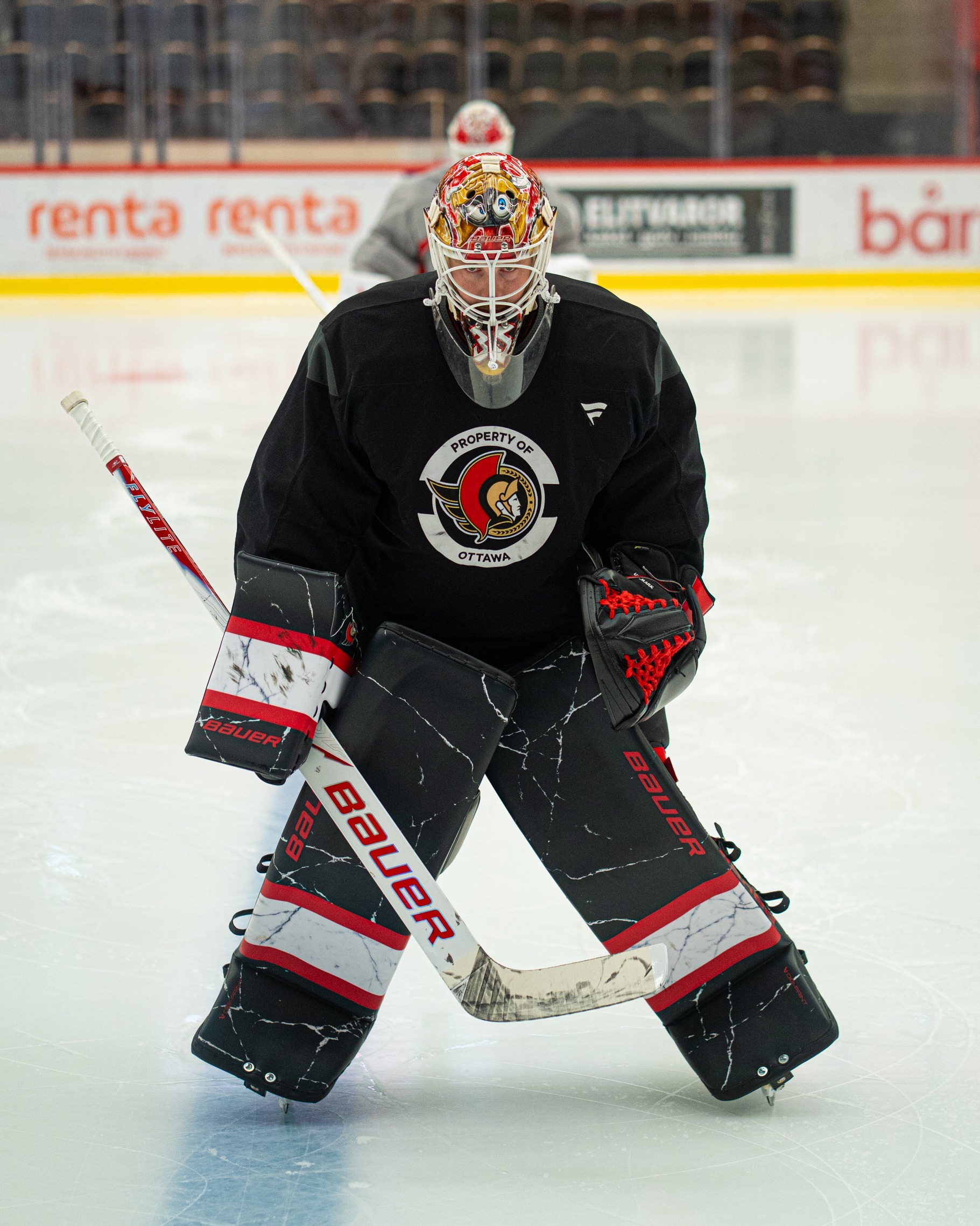 Linus Ullmark on ice in an Ottawa Senators practice jersey donning a black glove, blocker and pads with a large white stripe with smaller red stripes around it.