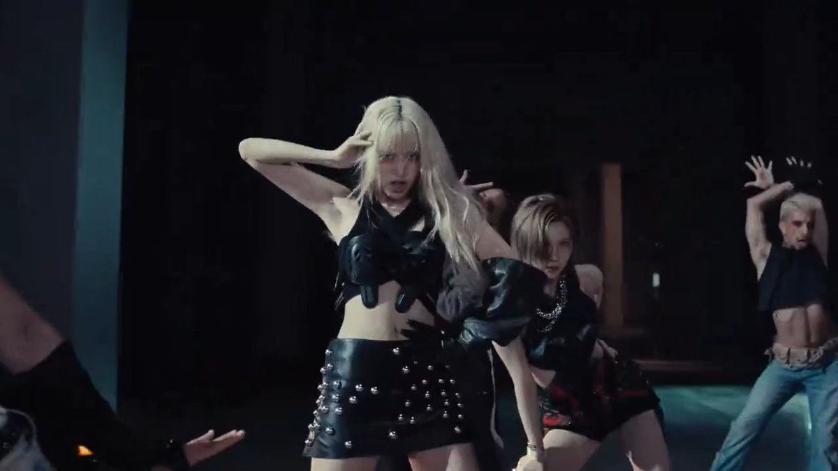A woman with long blonde hair in a black leather outfit and studded skirt dances in a dark, industrial setting, accompanied by backup dancers in the background.