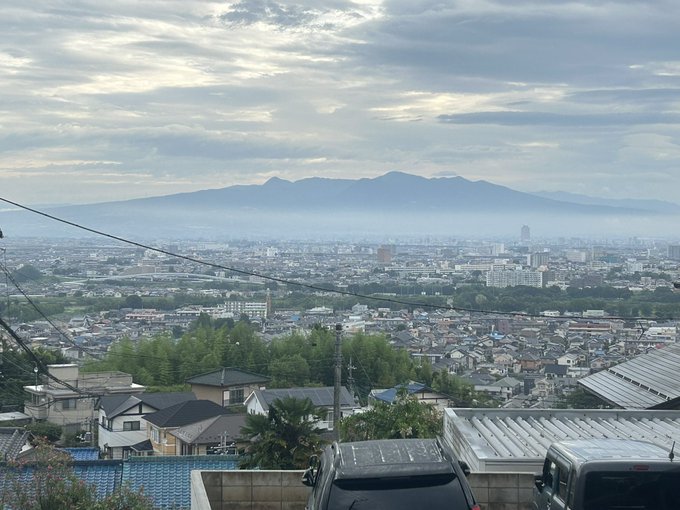The image shows a distant view of Mount Akagi (Akagiyama) under a cloudy sky. The mountain is clearly visible despite the overcast weather. In the foreground, there are rooftops of residential buildings and some greenery, indicating a suburban or urban setting. The post text mentions that although it is cloudy, Mount Akagi is clearly visible, and the author is sharing this view with those who couldn't return home. This context adds a sense of sharing and connection with the mountain's beauty.