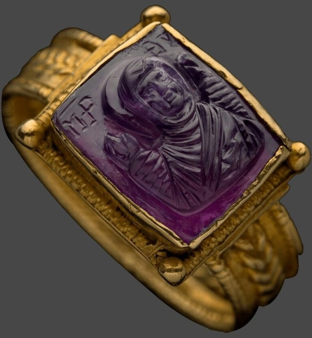 Ring; Byzantine; 11th century AD.

A rectangular bezel holds a carved purple stone showing the Virgin Mary at half length with hands raised in prayer (orans). Flanking letters include the standard Greek abbreviation for “Mother of God” (ΜΡ ΘΥ).