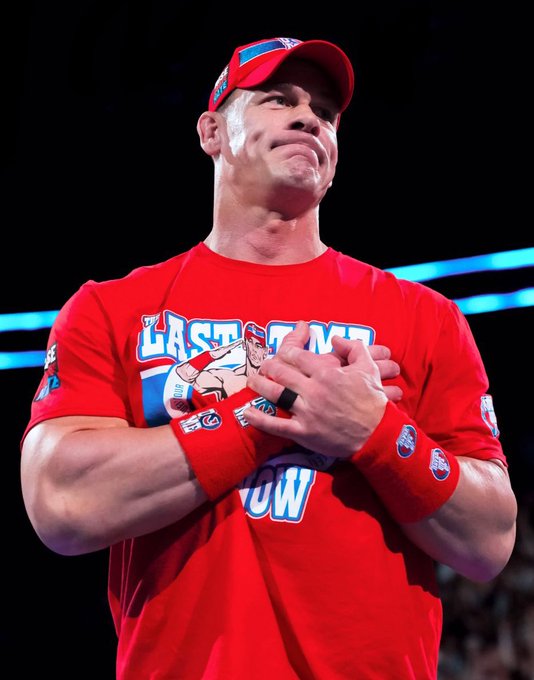 The image shows John Cena, a well-known professional wrestler, wearing a red "Last Time Ever" T-shirt and matching wristbands, with his hands clasped over his chest in a gesture of gratitude or farewell. He also wears a red cap, and the setting appears to be a WWE event with a dark background and blue lighting, suggesting an arena. The post text from Lucha Libre Online indicates Cena is leaving his final opponent choice to WWE for his last match in December, adding context to his pose as a poignant moment of retirement or farewell. No platform watermarks like Instagram, TikTok, or Xiaohongshu are visible.