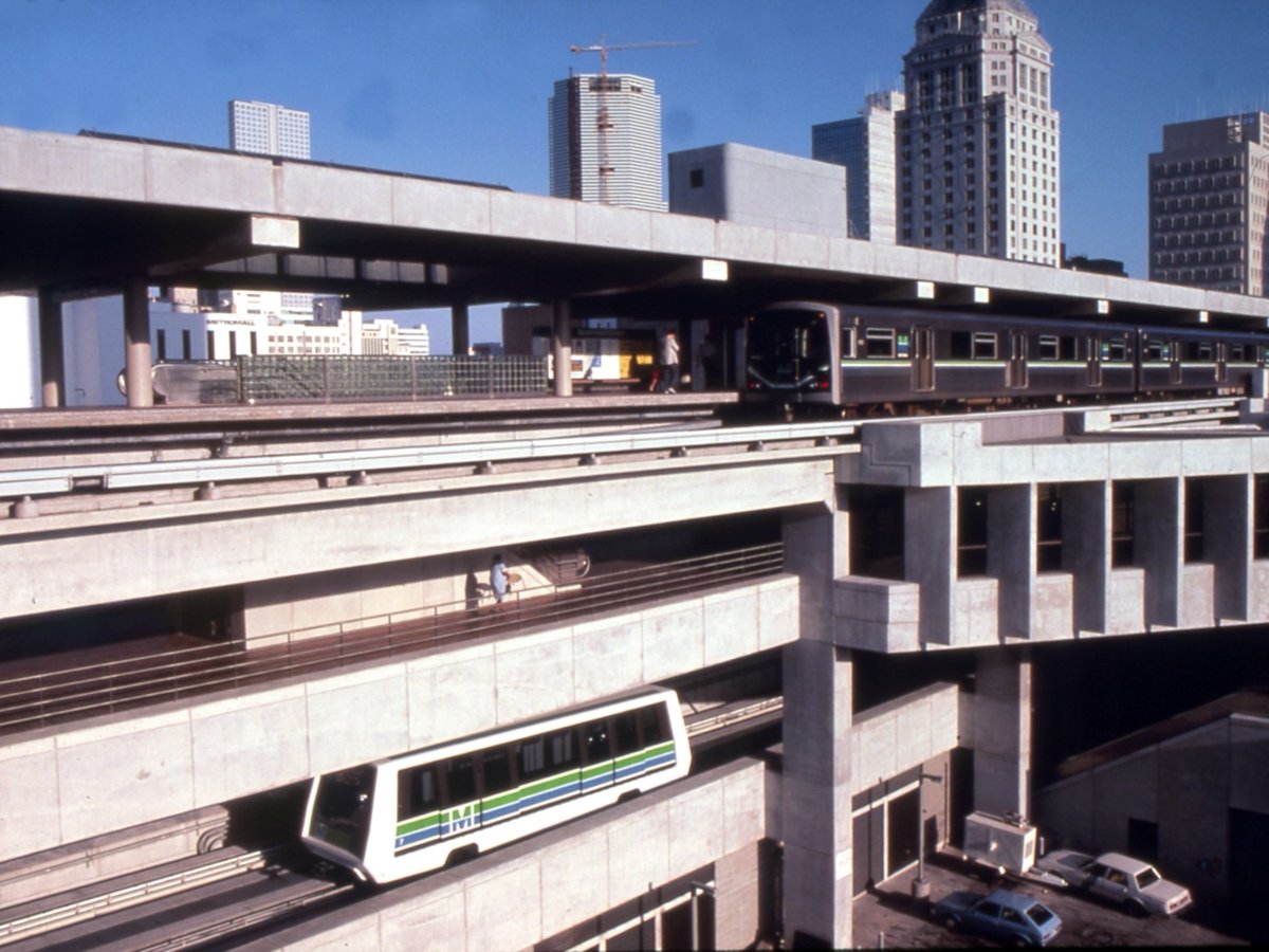 FLMemory's tweet image. Miami&apos;s Metrorail (top) and Metromover (bottom) running in close parallel, nestled against the Downtown Miami skyline at Government Center station, c. 1986. The vehicles are shown sporting their classic 1980s green-and-blue striped livery.