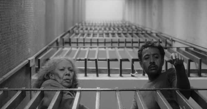The black-and-white image depicts two individuals leaning over a metal railing in a narrow, industrial staircase or corridor, with a long perspective of railings extending into the distance. The setting suggests a confined, urban environment, possibly symbolizing entrapment or tension, fitting the film "No Nos Moverán" (Pierre Saint-Martin Castellanos, 2024), a dark comedy fable about revenge and the legacy of the 1968 Tlatelolco massacre. The individuals’ expressions and poses hint at a dramatic or ironic moment, aligning with the film’s themes of reflection and historical tragedy. No platform watermarks are visible.
