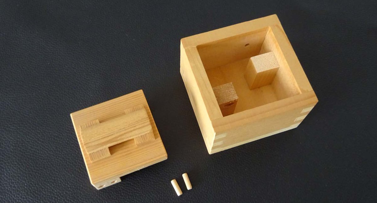 片桐厚氏がデザイン・制作したTrick BOX。取っ手のついた蓋を開けるのが目的。蓋はカタカタと動いて簡単に開きそうなのに開く気配はない。ふと思いついた方法を何度か試しているうちに突然開いて中から2本の小さなタボが飛び出してきた。このタボの働きで蓋を開けられなかった。IPP15の交換パズル。